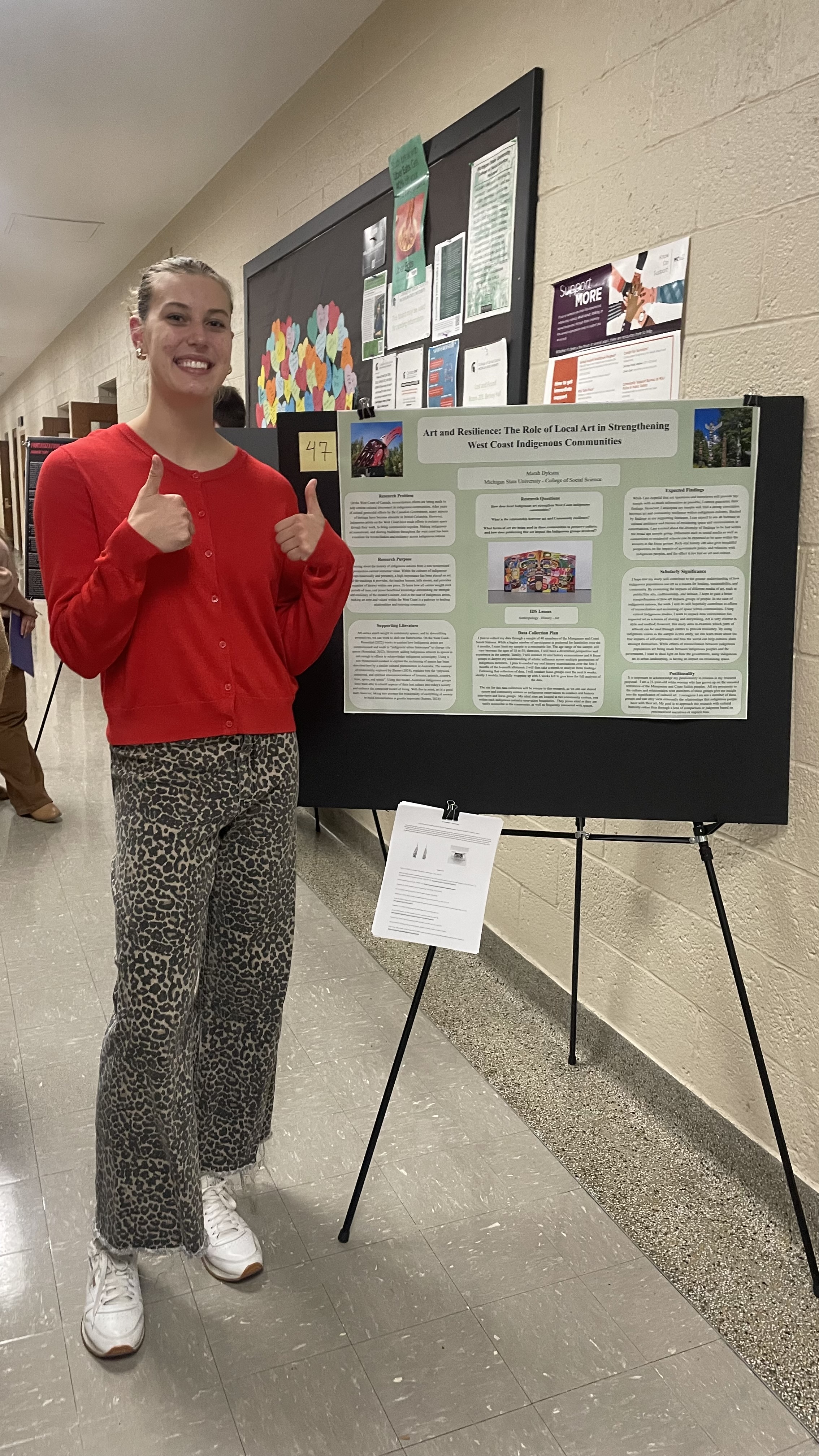 Photo of an MSU student smiling next to her poster presentation and giving two thumbs up
